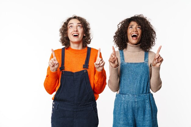 Retrato de alegre sorprendió a dos amigas multirraciales en monos de mezclilla aislados en la pared blanca apuntando a copyspace.