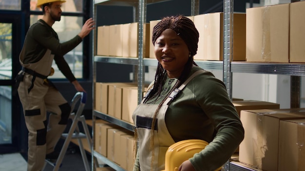 Retrato de un alegre trabajador de almacén afroamericano parado frente a estantes llenos de productos empaquetados en cajas de cartón listas para ser enviadas, feliz de trabajar en un ambiente profesional