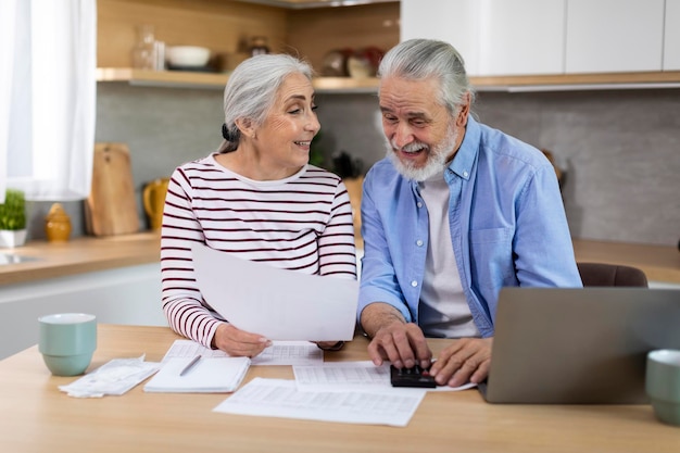 Retrato de alegres cónyuges ancianos sentados en la cocina y revisando documentos financieros