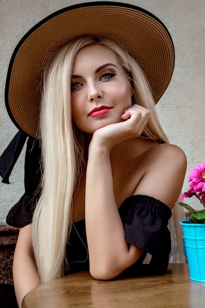 Retrato de alta moda de una mujer elegante con un elegante sombrero de paja y un vestido que se sienta en una cafetería en una mesa