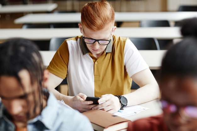 Retrato de alto ángulo de un joven pelirrojo que usa un teléfono inteligente en el escritorio en el aula universitaria