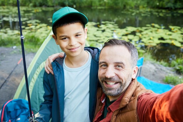 Retrato de alto ángulo de padre feliz tomando foto selfie con hijo mientras disfruta de un viaje de pesca juntos, espacio de copia