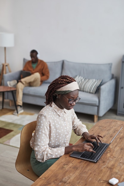 Retrato de alto ángulo vertical de joven afroamericana con laptop en el escritorio mientras trabaja desde casa en el acogedor interior del apartamento