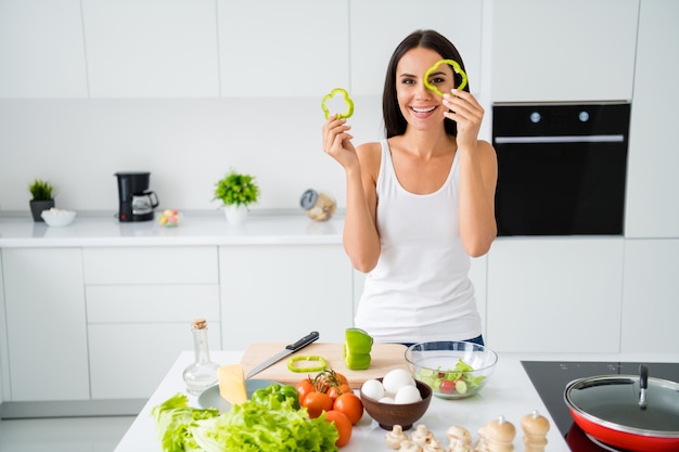 Retrato de ama de casa chica alegre positiva quiere ser chef preparar almuerzo vegetariano sostenga rodajas de pimienta imagina que es sus gafas usan camiseta blanca en casa de cocina en el interior