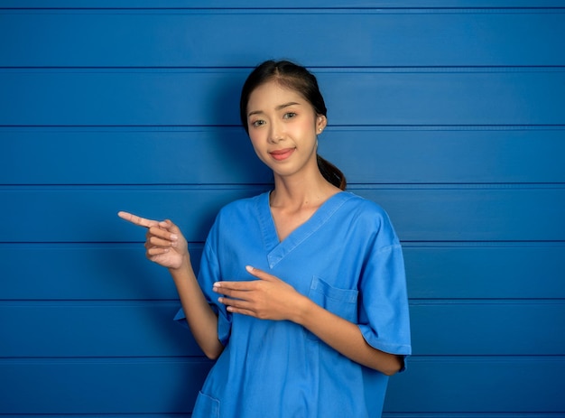 Retrato de una amable y confiada mujer asiática, doctora o enfermera, con traje de exfoliación azul, señalando con el dedo a la izquierda, mostrando el fondo azul de la pared de madera para publicidad sobre atención médica y médica