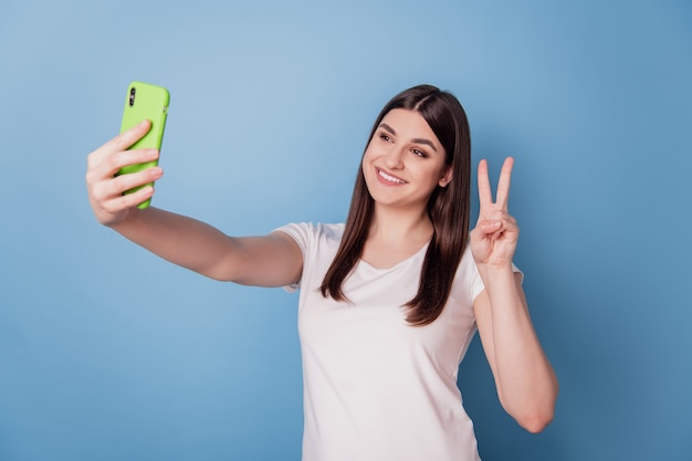 Retrato de amable dama positiva mantenga el teléfono hacer selfie mostrar signo v sobre fondo azul.