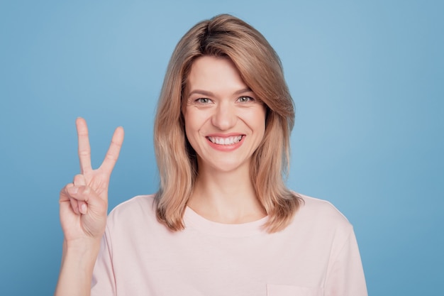 Retrato de amigable chica positiva dedos muestran gesto de paz sonrisa con dientes sobre fondo azul.