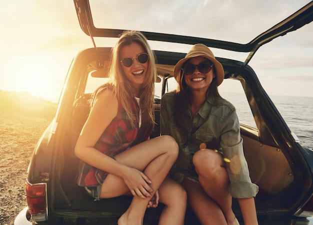 Retrato de amigas y baúl de automóvil en la playa de viaje por carretera o relajarse en vacaciones para la puesta de sol con una sonrisa Mujer al aire libre y amistad felicidad vacaciones en el océano o gafas de sol para el sol de verano juntos