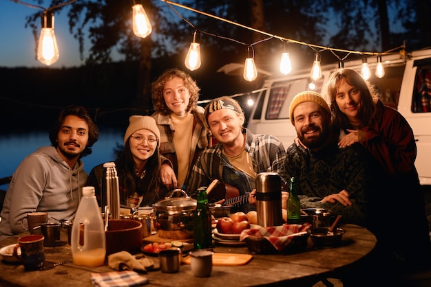 Retrato de amigos felices sonriendo a la cámara mientras está sentado en la mesa de la cena durante el campamento en el bosque