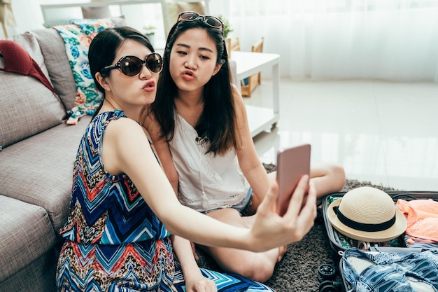 Retrato de amigos positivos de dos hermanas felices haciendo selfie en el teléfono inteligente faneca caras divertidas muecas alegría emociones estilo casual loco. las mujeres que se divierten preparan el equipaje para un viaje de lujo en verano