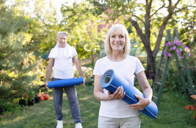 Retrato de una anciana activa posando con su esposo en el jardín sosteniendo colchonetas de yoga haciendo ejercicio juntos