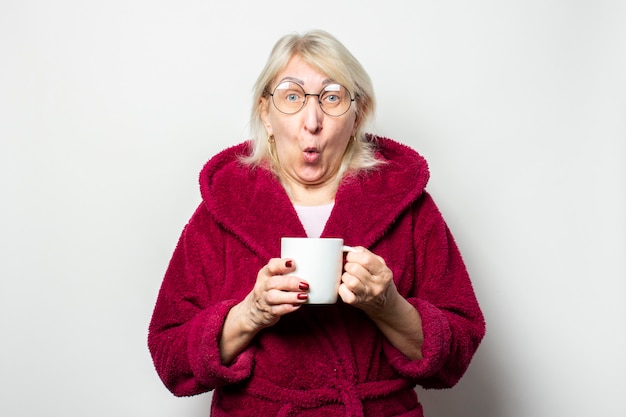 Retrato de una anciana amigable con una cara sorprendida en una bata todos los días y gafas sosteniendo una copa en la mano sobre una pared de luz aislada. Cara emocional Gesto de sorpresa, sorpresa