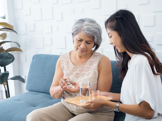 Retrato de una anciana asiática de cabello gris corto sostiene una píldora y un vaso de agua potable de una hija adulta joven que toma medicamentos diarios o suplementos vitamínicos para ancianos y médicos en el hogar