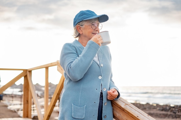 Retrato de una anciana atractiva y pensativa en vacaciones en el mar de pie en la playa disfrutando de una taza de té