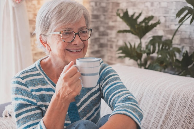 Retrato de una anciana de cabello gris sonriente caucásica sentada en un sofá en casa sosteniendo una taza de café