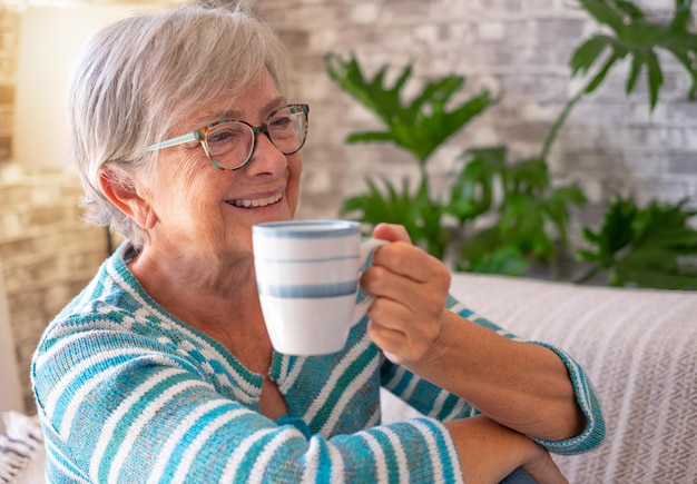 Retrato de una anciana caucásica sonriente sentada en un sofá en casa sosteniendo una taza de café Fondo de pared de ladrillo