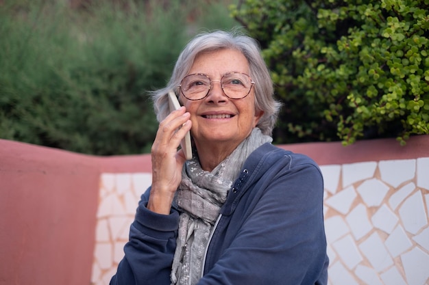 Retrato de una anciana feliz y relajada sentada al aire libre en un banco mientras habla por teléfono inteligente