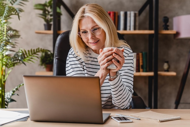 Retrato de una anciana feliz usando una laptop mientras bebe café en el lugar de trabajo