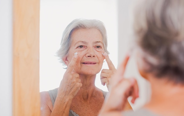 Retrato de una anciana hermosa de pelo gris aplica crema antienvejecimiento en la cara arrugada mirando el espejo cuidando el concepto de piel