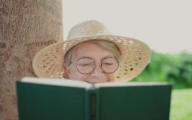 Retrato de una anciana relajada y atractiva con sombrero y anteojos sentada en el prado del parque público leyendo un libro dama caucásica disfrutando del tiempo libre y la jubilación