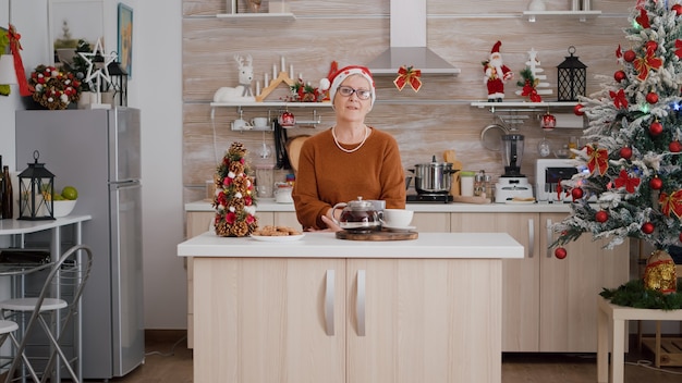 Retrato de anciana con sombrero de Navidad celebrando la temporada navideña en cocina decorada