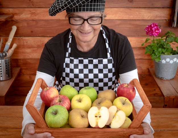 Retrato de una anciana sonriente con anteojos negros sosteniendo una canasta llena de manzanas frescas