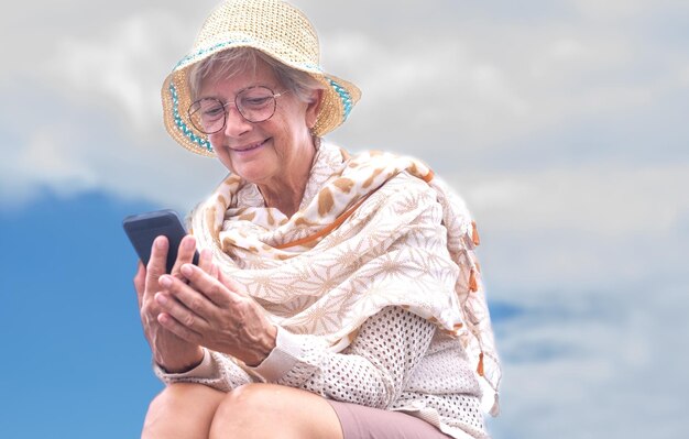 Retrato de una anciana sonriente con anteojos y sombrero de verano usando un teléfono móvil sentado al aire libre disfrutando de la libertad tecnológica y relajándose Espacio de copia del cielo nublado