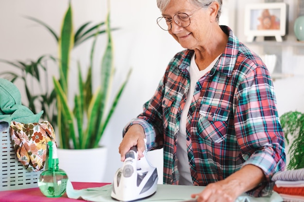Foto retrato de una anciana sonriente con camisa a cuadros y gafas planchando ropa en casa en una tabla de planchar