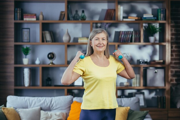 Retrato de una anciana sonriente en casa haciendo ejercicio y mirando la cámara sosteniendo pesas ejercicio matutino