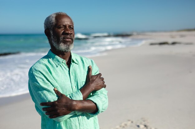 Retrato de un anciano afroamericano parado en la playa con cielo azul y mar de fondo, mirando a lo lejos con los brazos cruzados