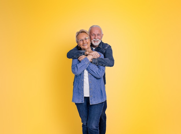 Retrato de un anciano barbudo amoroso romántico abrazando a su esposa por detrás y mirando a la cámara mientras posan juntos contra un fondo amarillo