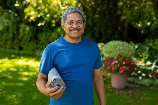 Foto retrato de un anciano birracial sonriente con ropa deportiva sosteniendo una alfombra de yoga contra las plantas en el patio