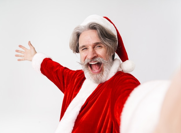 Retrato del anciano encantado con traje rojo de Santa Claus tomando retrato selfie y gritando aislado en blanco