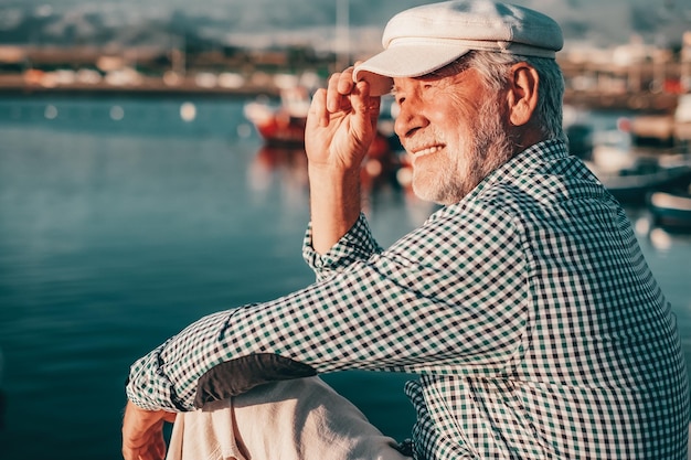 Retrato de un anciano feliz con camisa informal y sombrero sentado en el puerto marítimo al atardecer Relajado abuelo barbudo que disfruta de viajes de tiempo libre o jubilación