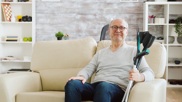 Retrato de anciano jubilado sonriendo a la cámara en un acogedor hogar de ancianos, tiene muletas junto al sofá