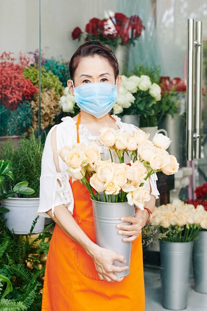 Retrato del anciano propietario de la tienda de flores de Asia en la máscara protectora de pie en la exhibición floral con un balde de rosas frescas shitw