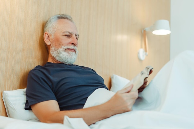 Retrato de un anciano sonriente de pelo gris acostado en la cama cubierto con una manta sosteniendo un libro leyendo en un elegante apartamento Un hombre maduro y guapo descansando en el hotel