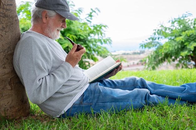 Retrato de un anciano sonriente sentado en el césped en un parque público con momentos relajados leyendo un libro Pipa de fumar masculina barbuda de ancianos caucásicos