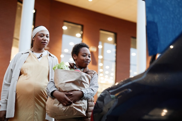 Retrato de ángulo bajo de adolescente afroamericano llevando comestibles al coche mientras ayuda a mamá a hacer compras, espacio de copia