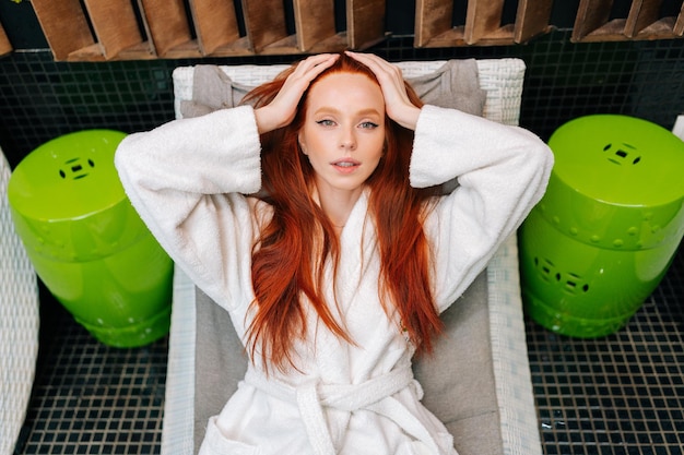 Retrato en ángulo alto de una joven alegre con albornoz blanco relajándose con los ojos cerrados tumbada en una tumbona después de un tratamiento de belleza en el salón de spa
