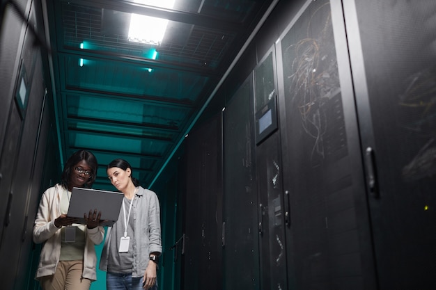 Retrato de ángulo bajo de dos mujeres jóvenes que usan la computadora portátil en la sala de servidores mientras configuran una red de supercomputadoras, espacio de copia