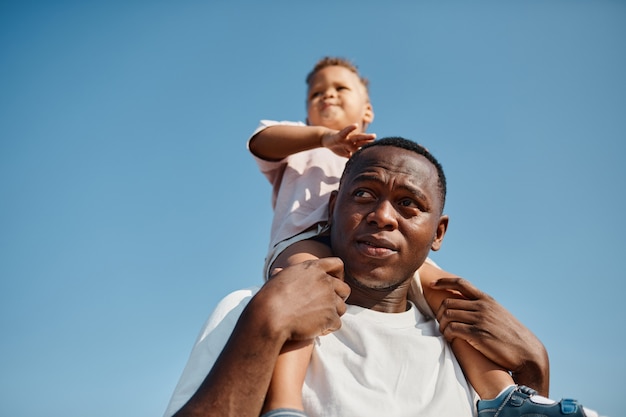 Retrato de ángulo bajo de feliz padre afroamericano con hijo en hombros contra el cielo azul mientras ...