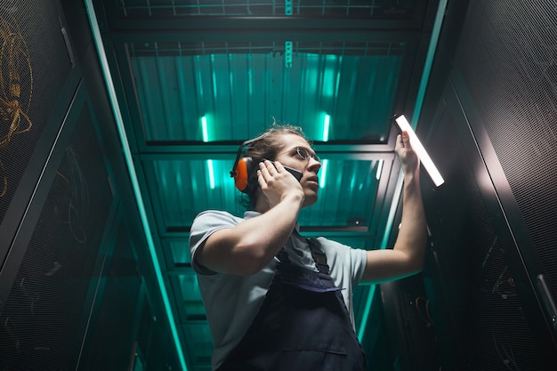 Retrato de ángulo bajo de ingeniero de red masculino hablando por teléfono inteligente en la sala de servidores mientras trabaja en el centro de datos, espacio de copia