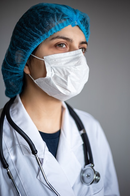 Retrato de ángulo de una joven doctora con máscara médica, bata y estetoscopio. Enfermera esperando al próximo paciente en un hospital para el control habitual. Prevención de coronavirus y concepto de visita al médico.