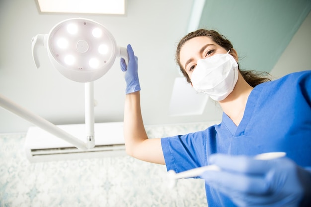 Retrato de ángulo bajo de mujer dentista con herramienta de ajuste de equipo de iluminación en clínica dental