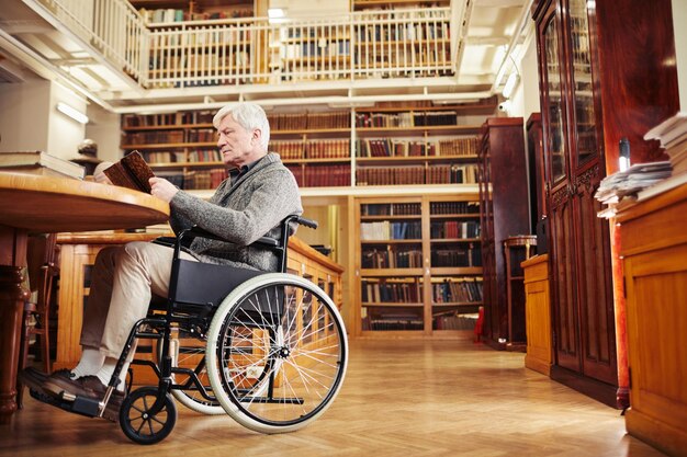 Retrato de ángulo bajo del retrato de un anciano que usa silla de ruedas y un libro de lectura en el espacio de copia interior de la biblioteca clásica