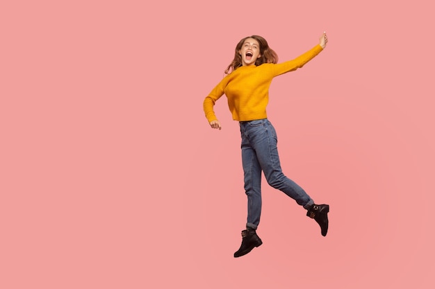 Foto retrato de una animada y enérgica jengibre con suéter y denim llena de entusiasmo saltando al aire o volando, gritando de felicidad. tiro de estudio interior aislado sobre fondo rosa, espacio de copia