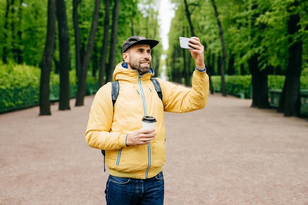 Retrato ao ar livre de bonitão com barba grossa vestindo amarelo anoraque e jeans segurando mochila café e smartphone fazendo selfie contra árvores verdes Turista satisfeito descansando no parque