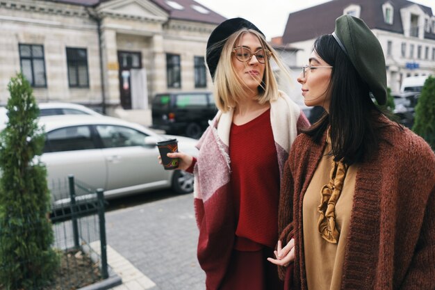 Retrato ao ar livre de duas amigas. garotas com roupas quentes casuais e óculos, passando pela cidade na estação fria e se divertindo na rua urbana. estilo de vida da cidade, conceito de amizade.