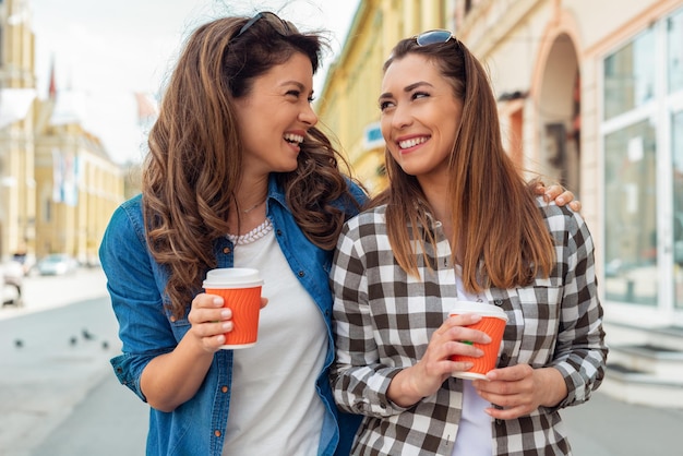 Retrato ao ar livre de duas meninas alegres bebendo café. Caminhando na cidade.